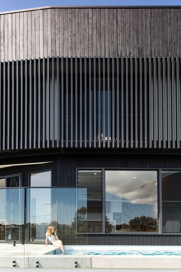 Contemporary multi-level residential facade with dark vertical cladding, a weathered timber upper volume, and a deep balcony screened by closely spaced black vertical fins. Large glazed doors and windows open toward a narrow swimming pool, with a glass balustrade along the pool terrace.