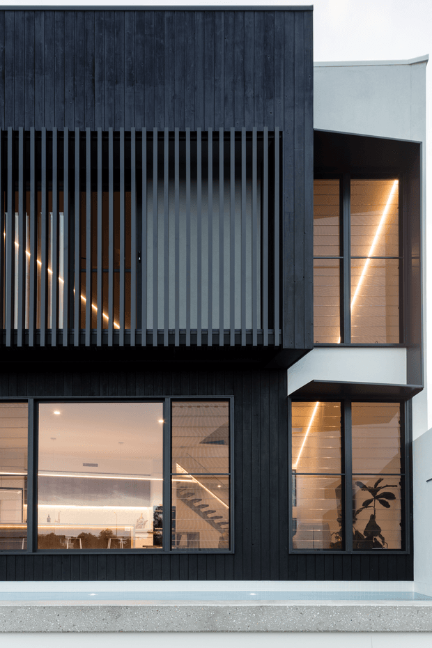 A contemporary multi-storey residential facade clad in dark vertical timber boards, with projecting upper-level volumes, black vertical screening, and large dark-framed glazed windows. Warm interior lighting is visible through the glazing, and a narrow pale-blue swimming pool runs along the base of the building.