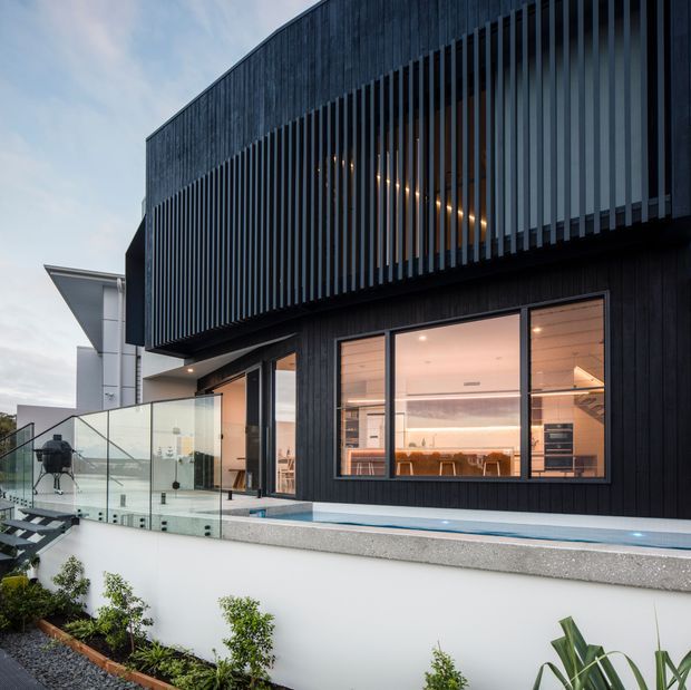 Exterior architectural photograph of a contemporary multi-level residence with dark vertical timber cladding and a prominent upper-storey louvred screen. Large glazed openings reveal a warm, modern kitchen and dining area. A glass balustrade, elevated terrace, narrow pool or water feature, and landscaped garden are visible in the foreground.