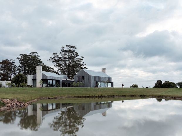 A contemporary rural residence composed of interconnected low-rise volumes, with dark vertical timber-like cladding, pale metal pitched roofs, extensive glazing, and a prominent light-toned chimney or vertical wall element. The house sits on an expansive grassy site beside a reflective pond, with mature trees and an overcast sky creating a quiet architectural landscape view.