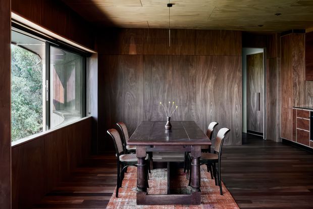 A dark, richly timber-clad dining room with a rectangular solid timber dining table, four upholstered chairs, and a small central vase. The room features extensive dark wood wall panelling and flooring, a patterned timber ceiling, large window glazing on the left, and a doorway leading to another space. Built-in timber cabinetry is visible at the right edge. The atmosphere is warm, moody, and refined, with strong mid-century-inspired detailing.