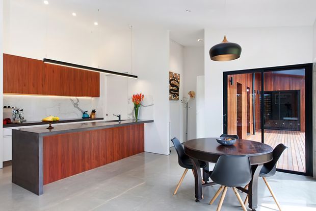 A bright, open-plan contemporary kitchen and dining area. The kitchen features warm timber cabinetry, a pale marble-look splashback, a dark island benchtop, integrated appliances, and a linear pendant light. A round dark timber dining table with modern chairs sits beside large black-framed glazing opening to a timber deck or courtyard.