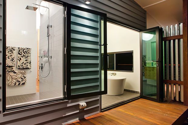 A contemporary bathroom viewed from a timber-floored adjoining space, featuring a glass-enclosed walk-in shower, freestanding white bathtub, wall-mounted rain shower, dark horizontal wall cladding, tiled bathroom surfaces, and extensive black-framed glazing and louvred screening. A green-toned vanity area is visible through the glass.