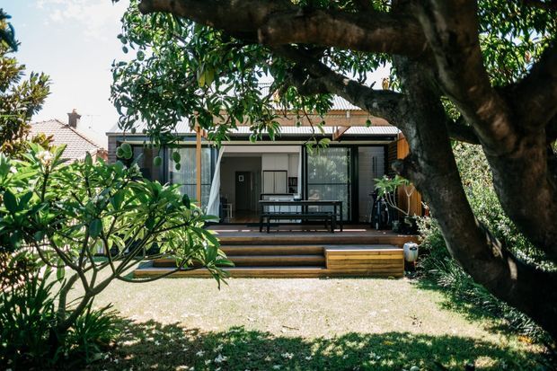 Exterior view of a contemporary single-storey home addition or pavilion set within a leafy garden. The building has broad glazed sliding doors, a covered timber deck with outdoor dining furniture, timber steps, vertical screening, and a low corrugated roof. A large established tree frames the foreground and partially obscures the facade.