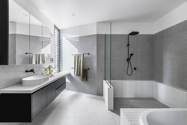 Contemporary bathroom with a spacious walk-in shower, freestanding bathtub partially visible in the foreground, wall-mounted matte black fixtures, large grey wall and floor tiles, a white small-format tiled shower base and bath surround, floating dark vanity cabinetry, white benchtop, frameless glass shower screen, and a large mirrored wall. Natural light enters through a louvred window, creating a clean, bright and refined atmosphere.