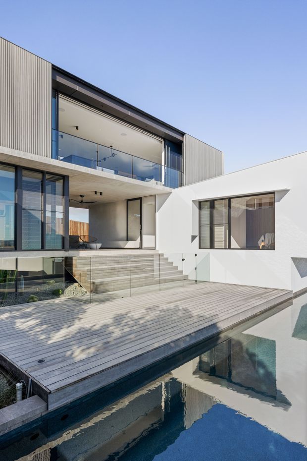 A contemporary multi-level residence arranged around a swimming pool and timber deck. The facade combines white rendered walls, dark-framed glazing, vertical ribbed cladding, exposed concrete, and a glass-balustraded upper terrace. Broad steps lead from the living level to the pool deck, with strong geometric forms and clean modern detailing.