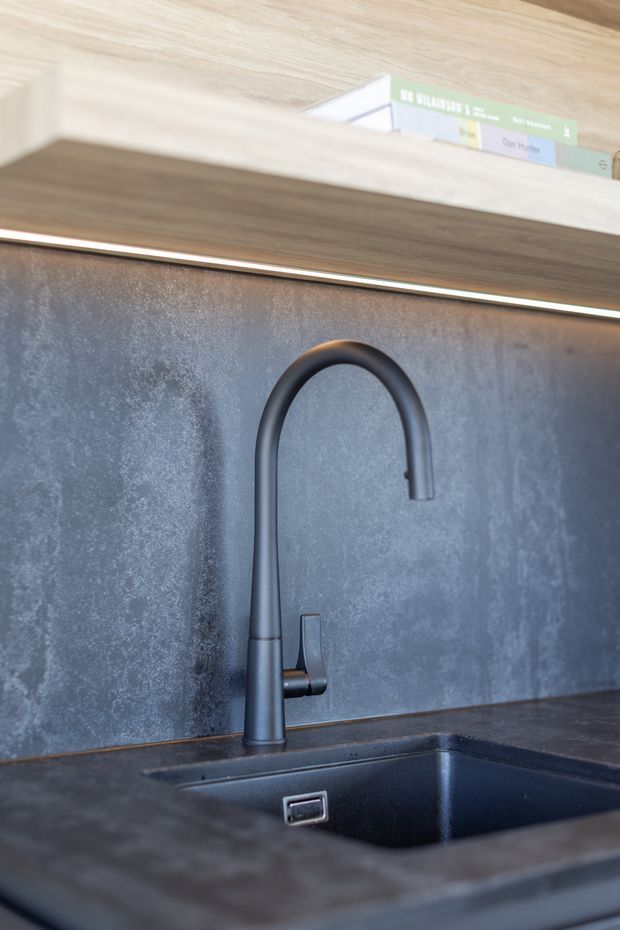 Close-up of a contemporary kitchen sink area featuring a matte black gooseneck mixer tap, integrated dark sink, dark stone-look benchtop, textured grey splashback, and warm timber cabinetry with linear under-cabinet lighting. A small shelf with books is visible above.