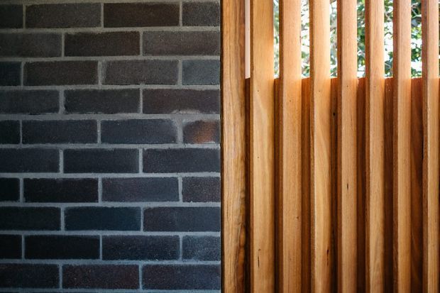 Close-up architectural detail showing a dark charcoal brick wall beside vertical warm timber slats, with narrow glimpses of greenery beyond the slatted screen. The image emphasizes contrasting masonry and timber textures rather than a complete room.