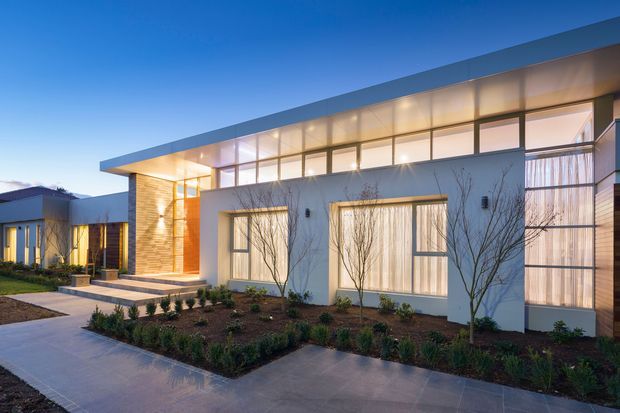 Twilight exterior view of a contemporary, low-profile multi-wing residence. The home features a long flat roof, extensive clerestory-style glazing, smooth light-coloured rendered walls, warm timber-toned vertical cladding at the entry, and carefully planted front garden beds with paved paths and steps.