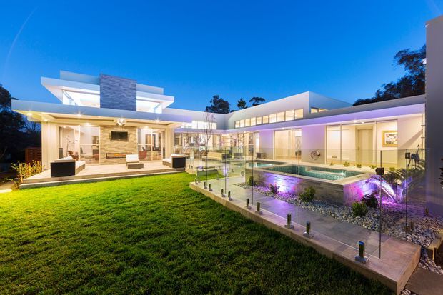 Twilight exterior view of a large contemporary flat-roofed residence arranged around a landscaped courtyard. The home features extensive floor-to-ceiling glazing, crisp white and light-coloured rendered walls, a projecting upper-level form, and a long illuminated wing. A raised rectangular swimming pool with glass balustrading, decorative gravel, and purple underwater and landscape lighting occupies the foreground beside a broad lawn.