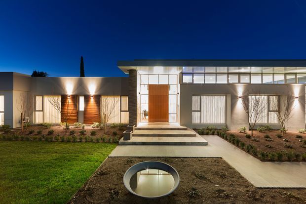 A wide dusk photograph of a low-slung contemporary residence with a strong horizontal composition. The facade combines pale rendered walls, extensive floor-to-ceiling glazing, a large timber pivot entry door, and illuminated exterior walls. Broad concrete steps lead to the entrance, with landscaped garden beds, lawn, and a circular recessed feature in the foreground. The roof appears flat and the architecture has clean, rectilinear lines.
