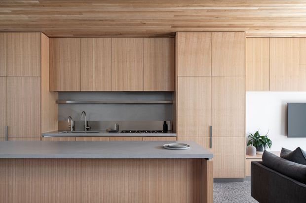 A refined open-plan kitchen with extensive light timber cabinetry and matching timber ceiling, a long grey island bench, integrated cooktop and sink, and a dark grey sofa visible in the adjoining living area. The kitchen has a clean, understated contemporary appearance with vertical fluted cabinetry on the island and full-height joinery.