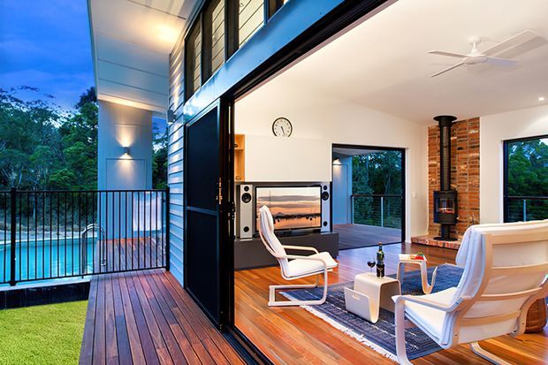 A contemporary open-plan living room opening onto a timber deck and balcony beside a swimming pool. The room features warm timber flooring, white walls, a large black-framed glazed opening, white lounge chairs, a media console, a brick feature wall with a freestanding wood-burning stove, and a ceiling fan. The exterior deck and pool are visible through the fully opened glazing.