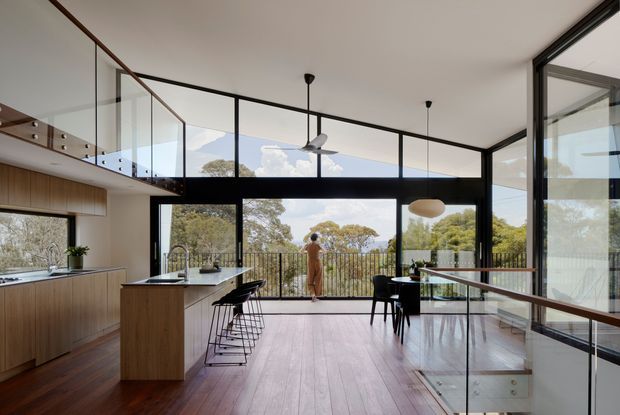 A bright, open-plan contemporary kitchen and dining area with a timber island, dark-framed floor-to-ceiling glazing, and a glass balustrade overlooking a lower level. Warm timber flooring and cabinetry contrast with black metal window frames, while a dining table and pendant lights sit beside the expansive outdoor view.