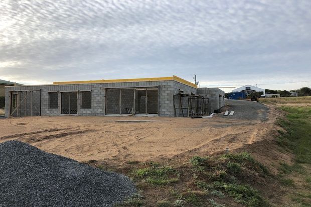 Single-storey building under construction, with exposed grey concrete block walls, unfinished openings and structural framing. The flat roofline has a bright yellow waterproofing or edge membrane, with a broad dirt construction yard, aggregate pile and construction materials in the foreground and surrounding site.