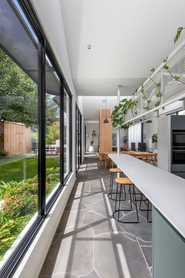Bright, elongated open-plan kitchen and dining space with floor-to-ceiling black-framed glazing along one side, opening views to a landscaped garden and outdoor dining area. A long pale island with timber stools dominates the foreground, with a dining table beyond, timber feature cabinetry, integrated dark appliances, and a white ceiling. A white perforated mesh balustrade with trailing greenery overlooks the room, suggesting a mezzanine or upper-level edge. The floor has large irregular grey stone-like slabs with light grout lines. The design is clean, contemporary, and strongly connected to the garden.