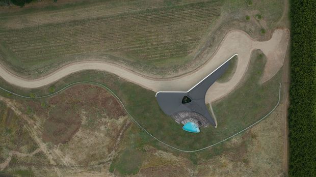 Aerial view of a sculptural, organically shaped building set within a broad rural landscape. The structure has a dark, three-winged or triangular roof form, curved pale paths or driveways around it, and a small bright turquoise pool or water feature near the lower edge. Surrounding fields and planted areas dominate the image.