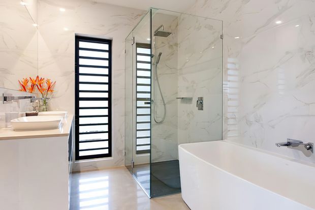 Bright contemporary bathroom with white marble-look wall tiles, a freestanding white bathtub, frameless glass shower enclosure, wall-mounted rain shower, floating vanity with vessel basin, and black-framed horizontal louvre windows. Orange flowers add a small accent beside the vanity.