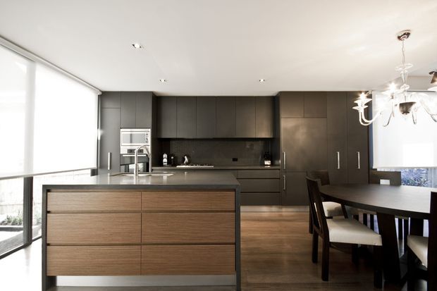 Contemporary open-plan kitchen and dining area with a large wood-fronted island, dark charcoal cabinetry, integrated stainless-steel appliances, dark splashback, and warm timber flooring. Full-height glazing brings in abundant daylight, while a dining table and chandelier occupy the adjacent area.