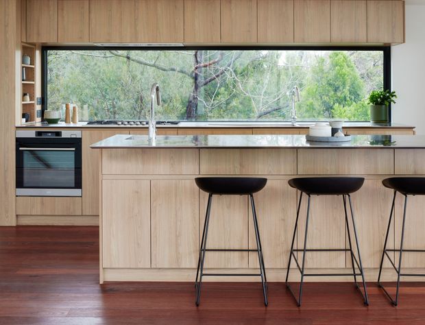 A contemporary kitchen with extensive light timber cabinetry, a long stone island with integrated sink and tap, and three black bar stools. A wide horizontal window provides a view to leafy vegetation, while dark reddish timber flooring adds warmth and contrast. Built-in appliances and open shelving are visible along the left wall.