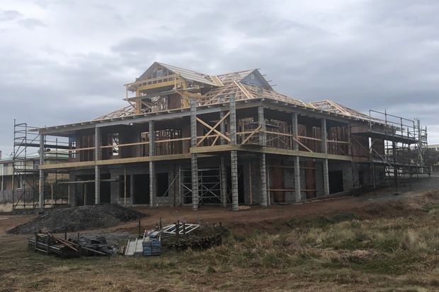 A multi-storey timber-framed building under construction, with exposed structural framing, partially completed pitched and gabled roof forms, scaffolding, and unfinished concrete or masonry lower-level walls. The building sits on a rough construction site under an overcast sky.