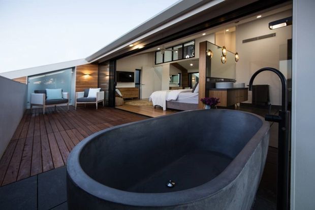 A contemporary indoor-outdoor retreat viewed from a dark freestanding bathtub on a timber deck. The open glazed room contains a bedroom with a bed, a compact vanity or bathroom zone, built-in cabinetry, pendant lights and recessed ceiling lighting. Sliding glass openings connect the interior to a furnished outdoor terrace, creating a strong sense of continuity between bathing, sleeping and outdoor living areas.