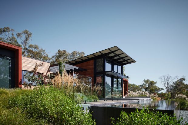 Contemporary single-storey residence set within a landscaped garden, featuring strong horizontal and vertical volumes, dark framed glazing, warm timber cladding, and a projecting flat canopy. A reflective swimming pool occupies the foreground beside the house, with native-style planting surrounding the terrace.