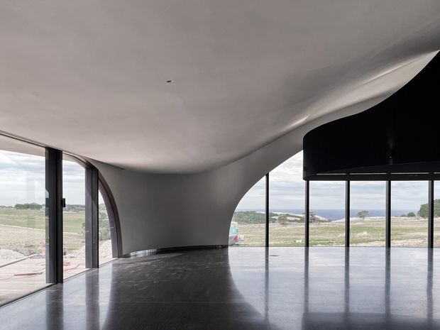An expansive, largely empty modern interior with a dramatic sculptural white curved ceiling and integrated curved wall forms. Full-height dark-framed glazing wraps the space, opening views toward a coastal or rural landscape. The polished dark floor strongly reflects the architecture and windows, while a large black curved overhead element occupies the right side.