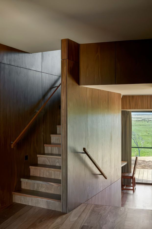 Interior view of a contemporary timber-lined stairway and landing. The staircase has open risers or slim-profile steps, dark wood wall panelling, integrated timber handrails, and a large central timber-clad partition with geometric panel joints. Beyond the partition is a dining area with a table and chairs, full-height curtains, and glazing overlooking a green rural landscape. The room is warmly lit with a refined, low-contrast material palette.
