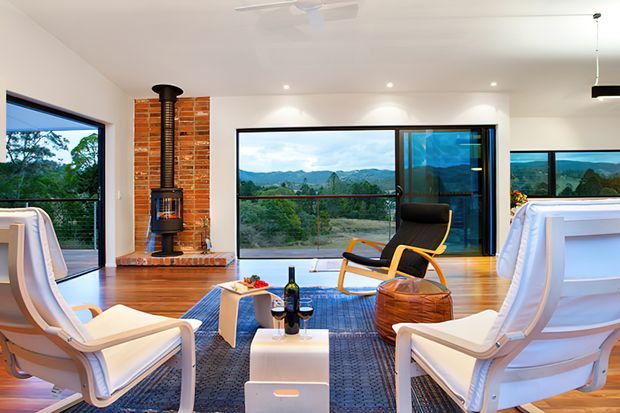 Bright, contemporary living room with warm timber flooring, large floor-to-ceiling glazing, and expansive views of a lush mountainous landscape. The room features a central brick fireplace with a black freestanding wood stove, white lounge chairs, a black sling chair, a blue area rug, and a low white coffee table. A ceiling fan and recessed lighting complete the relaxed open-plan interior.