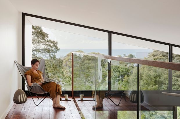 A bright, minimalist living area with full-height black-framed glazing overlooking leafy vegetation and a distant coastal horizon. A person sits in a suspended sling chair beside a small side table, while a glass balustrade encloses an upper-level edge or stair opening. The room features white walls, a dark timber floor, slim dark window frames, and abundant natural light.