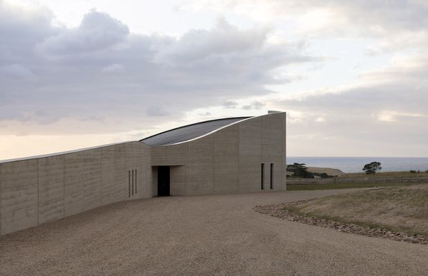 A low-profile contemporary coastal building set in an open gravel landscape. The monolithic pale concrete facade has strong horizontal board-formed texture, narrow vertical windows, and a dramatic curved roofline that rises toward one end. A dark recessed entry interrupts the otherwise solid wall, while the ocean and distant shoreline are visible beyond the site under an overcast sky.