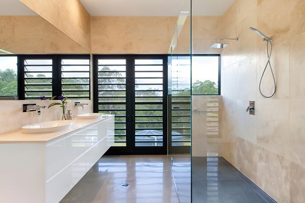 Bright contemporary bathroom with a double vanity, glossy white built-in cabinetry, large frameless glass shower enclosure, wall-mounted rain shower, and expansive black-framed louvred windows. The room features polished light grey concrete or stone flooring and warm beige textured wall finishes, with strong natural light and leafy suburban views beyond the windows.