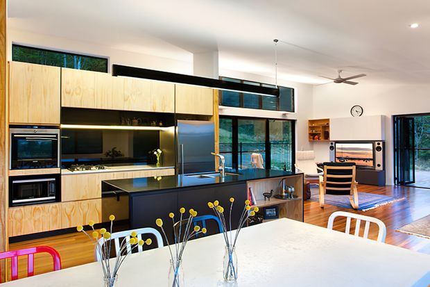 Open-plan contemporary kitchen and dining area with extensive light timber cabinetry, a dark island bench, integrated appliances, black-framed glazing, and timber flooring. The room opens toward a living area with a television and built-in shelving, while colourful dining chairs and small floral arrangements add playful accents.