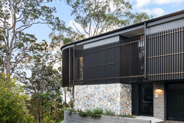 Exterior view of a contemporary elevated residence set among mature trees. The building features a long low-pitched roofline, dark vertical screening and cladding, extensive glazing, a textured irregular-stone wall, and a grey concrete-block base with planted edging. The projecting upper volume and screened balcony or walkway create a strong horizontal, modern composition.