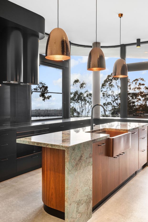 A contemporary kitchen with a large waterfall-edge island, dark built-in cabinetry, warm timber drawer fronts, and a polished green-toned stone benchtop. Three hammered metallic pendant lights hang above the island, while expansive black-framed glazing provides views of trees and sky. A sculptural black rangehood and curved island end add strong architectural character.