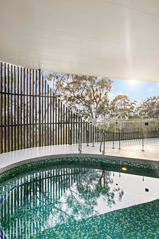 A covered outdoor pool area with a curved swimming pool finished in small green and blue mosaic tiles. The space has a broad white ceiling, dark vertical slatted screening, frameless glass pool fencing with black fittings, stainless steel handrails, and views of mature trees beyond. The reflective water and filtered daylight create a clean, tranquil atmosphere.