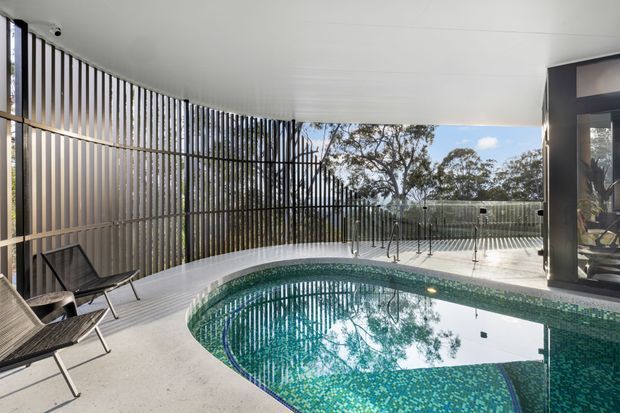 A covered outdoor pool area with a curved swimming pool, pale tiled decking, and a dramatic curved vertical-slat screen enclosing the space. Glazing and a dark-framed opening connect the pool area to the interior, while surrounding trees are visible beyond the enclosure. The design is contemporary and sculptural, with a calm, resort-like atmosphere.