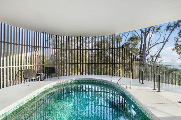 A bright covered pool area with a curved swimming pool finished in small green and blue mosaic tiles. The space is enclosed by tall vertical timber-look screening and clear glass balustrades, creating filtered views through surrounding trees toward a distant urban or coastal landscape. Minimal outdoor seating and stainless-steel pool handrails are visible on a pale tiled or concrete deck.