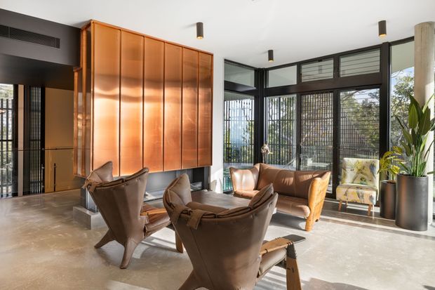 A sophisticated open-plan living room or lounge with a polished concrete floor, a dramatic floor-to-ceiling copper-toned timber feature wall and concealed cabinetry, black-framed floor-to-ceiling glazing, and sculptural brown leather lounge chairs arranged around a low coffee table. Natural light fills the room, with indoor plants and a warm neutral palette adding softness to the contemporary interior.