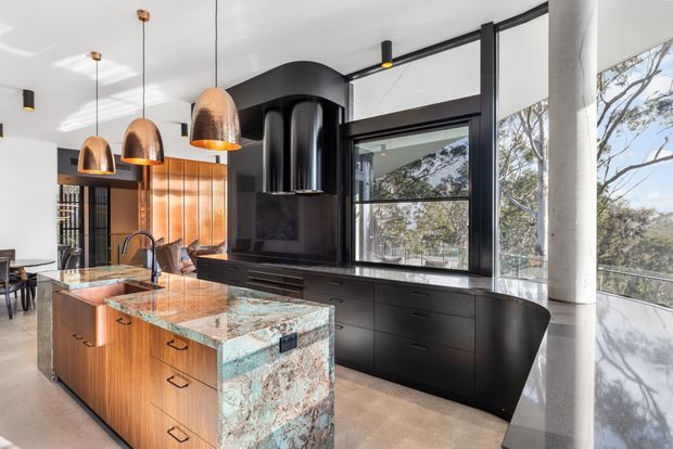A high-end contemporary kitchen with a large timber island and dramatic turquoise-veined stone end panel, black integrated cabinetry, a sculptural black rangehood, and expansive glazed windows. Copper pendant lights hang above the island, while a curved white structural column and views of trees outside add architectural character.