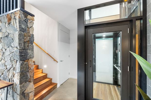 Bright contemporary entryway with a black-framed glazed front door and transom, opening toward a timber-floored interior. A timber staircase with a simple timber handrail rises beside a prominent rough natural-stone feature wall, with white walls, concealed storage doors, and polished concrete flooring. Indoor planting is visible at the right edge.