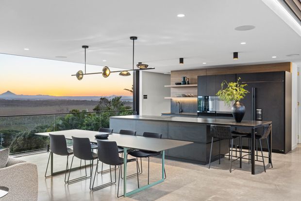 Contemporary open-plan kitchen and dining area with a large dark island, integrated cabinetry, pendant lighting, and a glass dining table with modern chairs. Full-height glazing opens to an expansive landscape view, while neutral stone-look flooring and a clean white ceiling create a bright, refined interior.