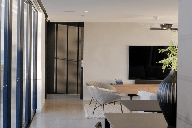 Bright contemporary living room with full-height glazed doors or windows along the left side, a dark framed internal door, pale smooth walls, and a large wall-mounted television above a timber media console. The room includes sculptural lounge chairs, nested dark coffee tables, a textured light rug, a large ribbed black vase, and a ceiling fan. The space has a clean, refined and airy appearance with strong indoor-outdoor glazing.