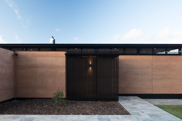 Single-storey contemporary building facade viewed from the front, with warm brown panelled walls, a dark low-profile roof edge, and a prominent black vertical slatted screen enclosing the entry. A pale paved walkway crosses a gravelled garden bed toward the entrance, with exposed dark structural framing and glazing visible along the upper facade.