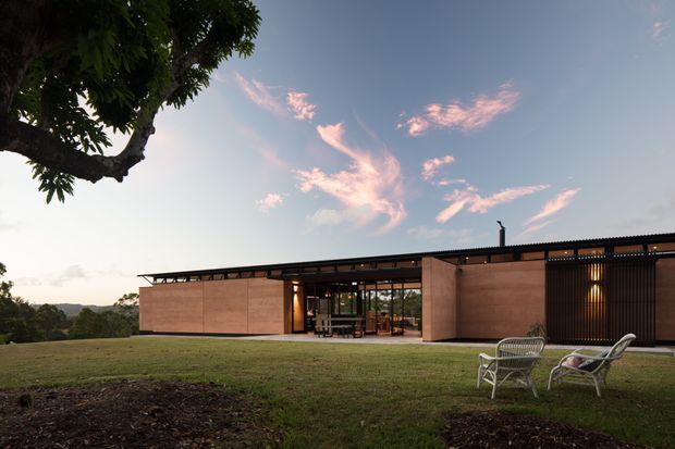 Single-storey contemporary residence set in a landscaped, elevated suburban or rural site, photographed at dusk. The long, low building has warm earthy rendered or masonry walls, a shallow dark corrugated roof, extensive glazed openings at the central living area, and a dark vertical-screened section at the right. Outdoor lounge chairs sit on the lawn beneath a mature tree, with distant terrain visible beyond.