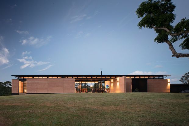A single-storey, low-profile modern building viewed from a broad lawn at dusk. The facade combines warm timber or wood-toned wall panels, extensive glass at the central entrance, dark recessed openings, and a very thin projecting flat roof with linear lighting beneath. A mature tree frames the right side, while the wide grass foreground and open sky emphasize the horizontal architectural composition.