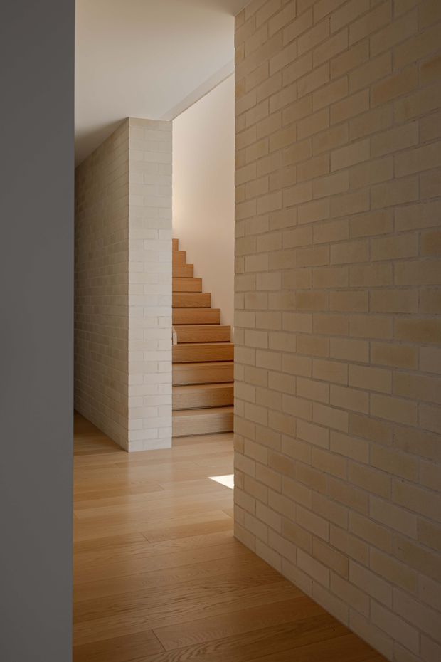 A warm, minimalist interior view through a narrow passage toward a straight timber staircase. Pale exposed brick walls form the circulation space, while light timber flooring and stair treads create a restrained natural palette. Soft daylight enters from the right, emphasizing the geometric wall planes and stair opening.