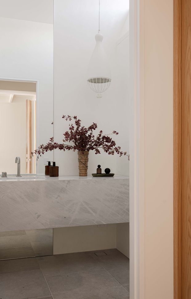 A refined minimalist bathroom vanity viewed through a doorway. The room features a large pale veined stone or marble-look floating vanity with an integrated basin, brushed metal tapware, mirrored wall panels, grey tiled flooring, warm timber door framing, and a sculptural white pendant light. Decorative branches in a textured vessel and small dark accessories add restrained contrast.