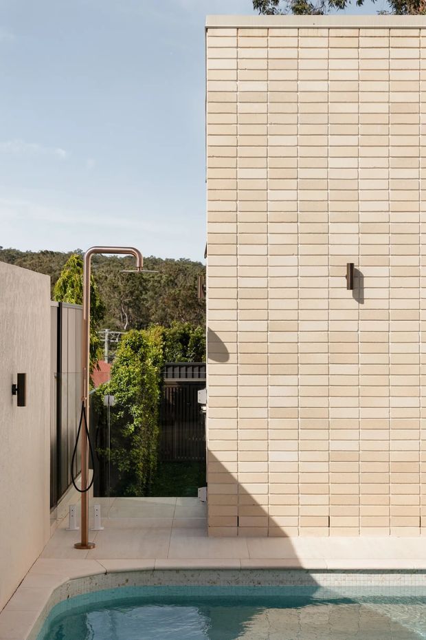 A compact outdoor poolside courtyard with a pale cream brick wall, tiled paving, a freestanding copper-toned outdoor shower, and a small swimming pool in the foreground. A timber privacy fence, garden vegetation, and neighbouring landscape are visible beyond the courtyard. The scene has clean contemporary detailing and warm natural daylight.